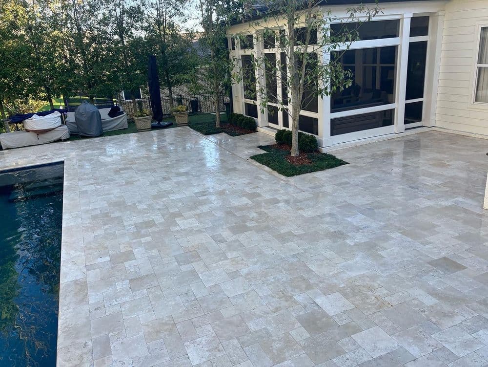 Elegant outdoor patio with travertine tiles, featuring a pool, greenery, and a screened porch.
