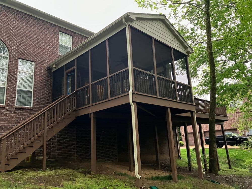 Screened-in porch on a brick house with stairs, surrounded by trees and greenery.