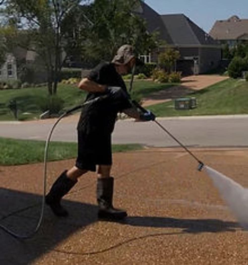 Man pressure washing a driveway on a sunny day, wearing gloves and rubber boots.