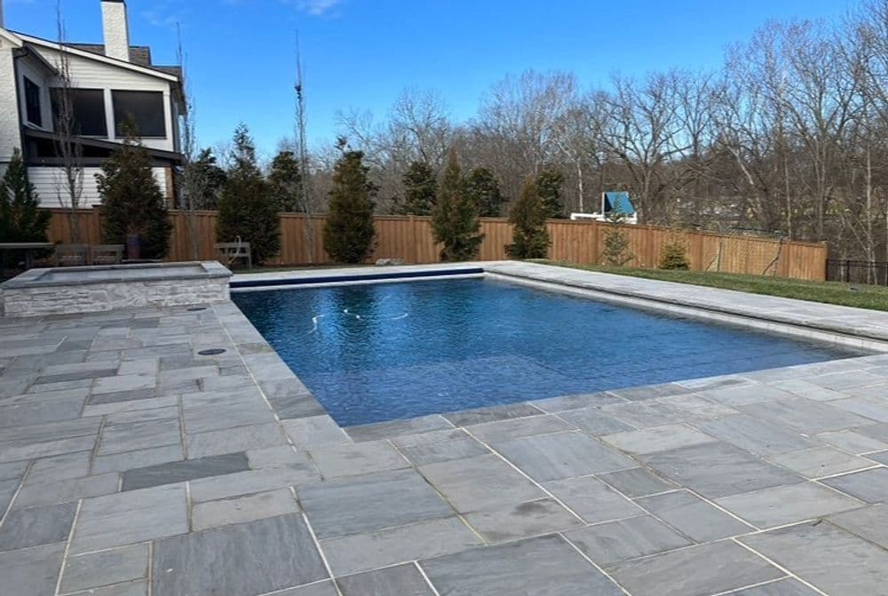 Modern backyard swimming pool with gray stone patio, wooden fence, and trees in the background.