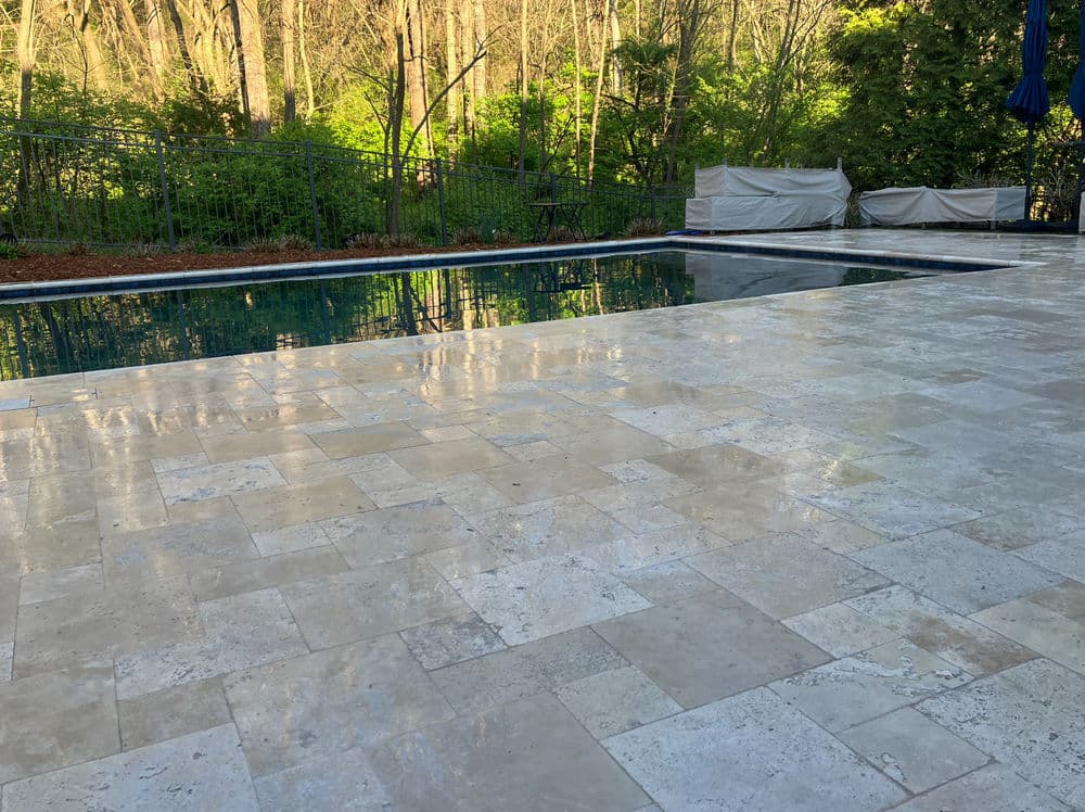 Serene backyard pool area with sunlit travertine patio and lush green trees in the background.