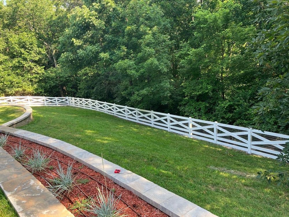 White wooden fence curving along a green lawn, surrounded by lush trees and landscaping.