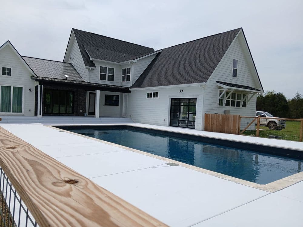 Modern farmhouse with a pool surrounded by a wooden deck and lush green landscape.