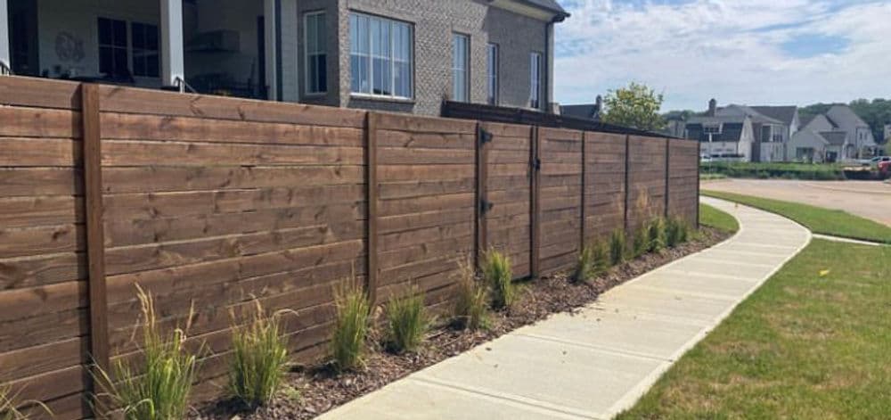 Modern wooden fence lined with ornamental grass beside a concrete pathway and a residential home.