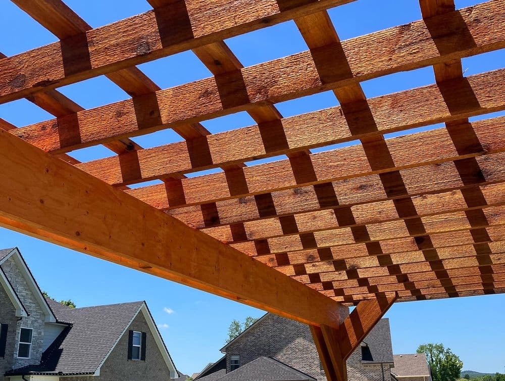 Wooden pergola structure with open lattice against a clear blue sky and residential houses.
