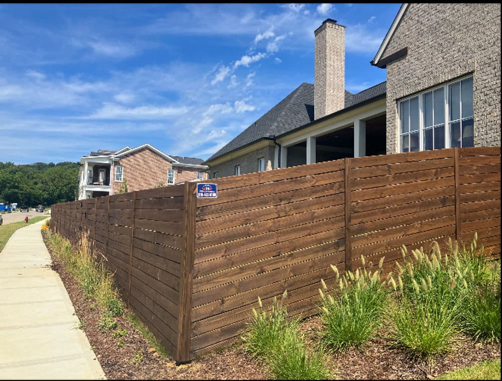 Modern wooden privacy fence with a house and blue sky background. Well-maintained landscaping.