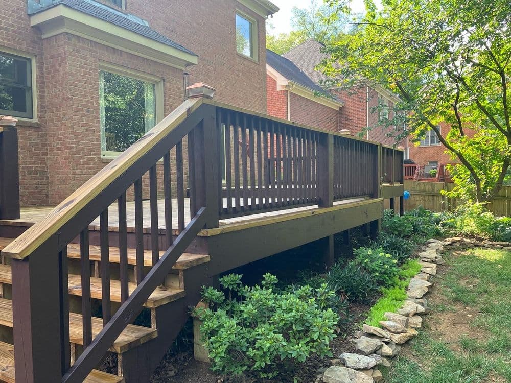Deck with wooden railing and steps in a backyard, surrounded by greenery and stone path.