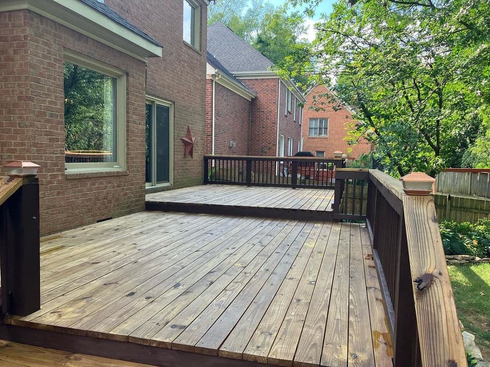 Wooden deck in a backyard with greenery, adjacent brick homes and a clear blue sky.