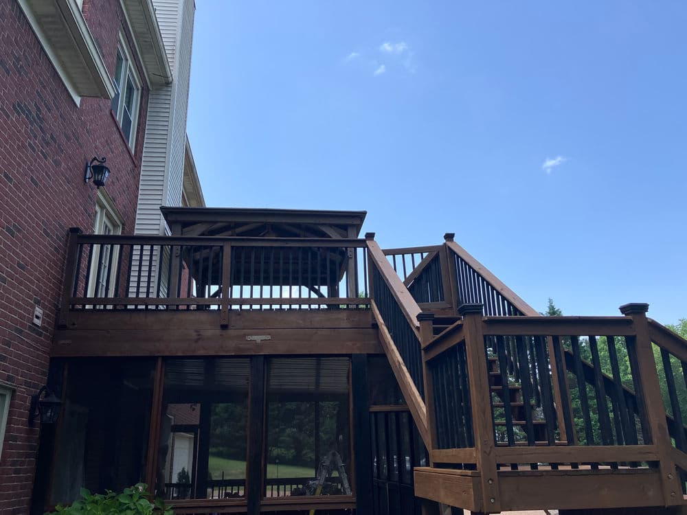 Two-tier wooden deck with gazebo and stairs, surrounded by greenery and blue sky.