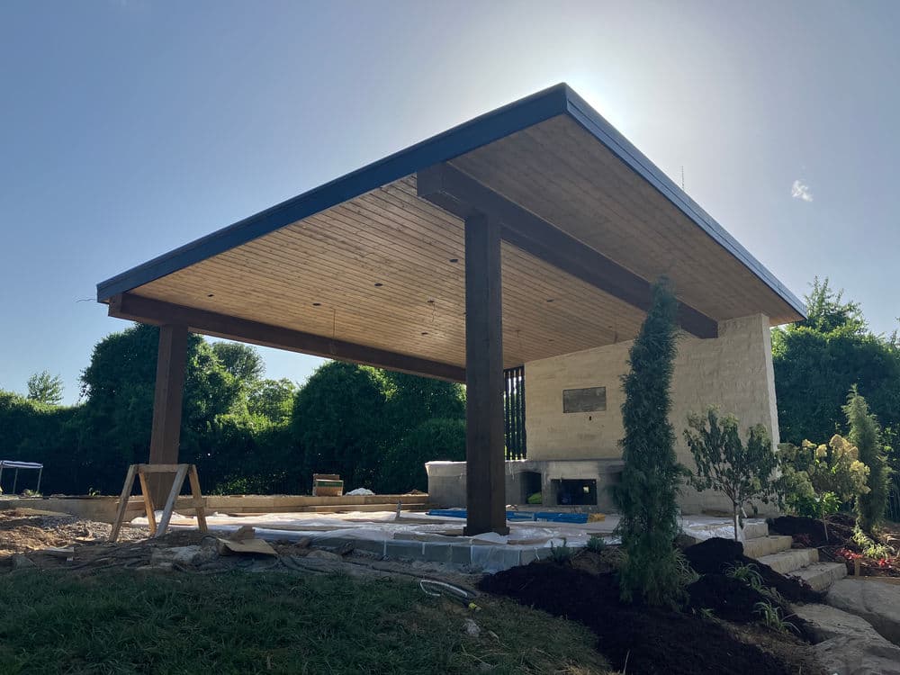 Modern outdoor pavilion with a wooden ceiling and stone walls, surrounded by green landscaping.