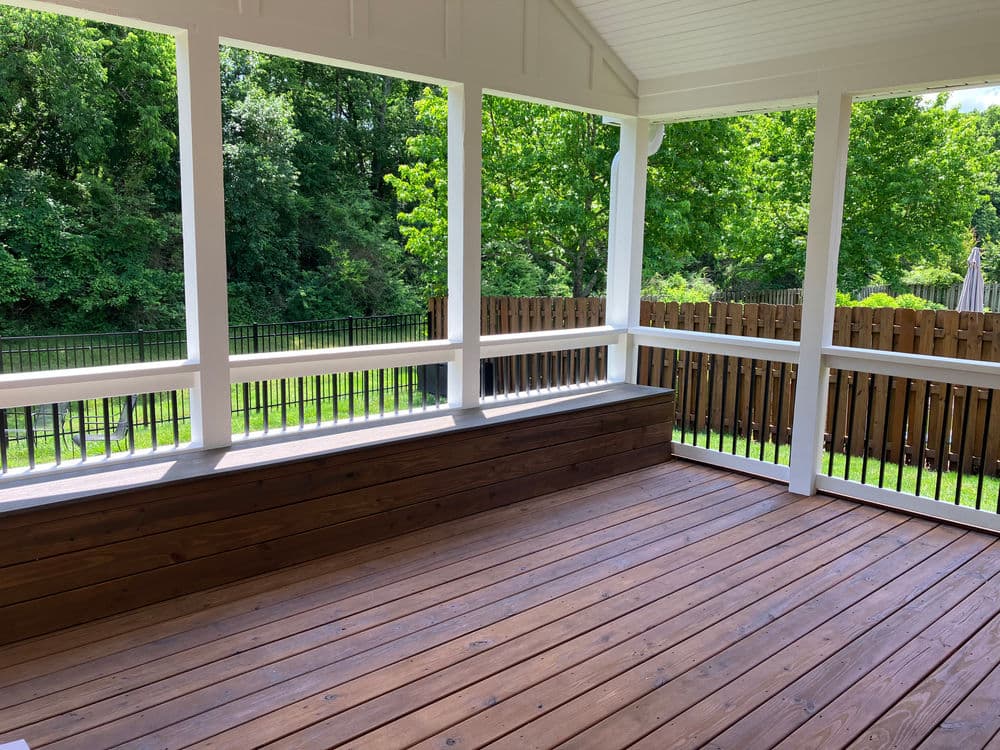 Spacious outdoor porch with wooden flooring and views of green trees and fenced yard.
