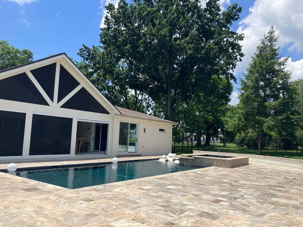 Modern house with pool and stone patio, surrounded by green trees and blue sky.