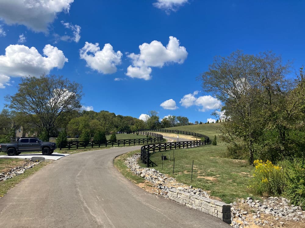 Winding road through green landscape under a blue sky with fluffy clouds and trees.