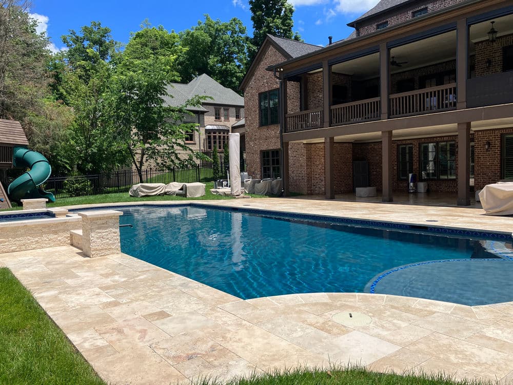 Backyard pool with slide, lounge area, and brick house, surrounded by lush greenery.