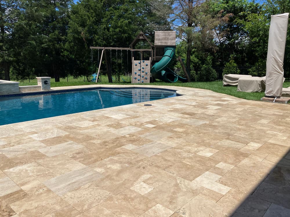Swimming pool with stone patio, slide, swing set, and lush greenery in the background.
