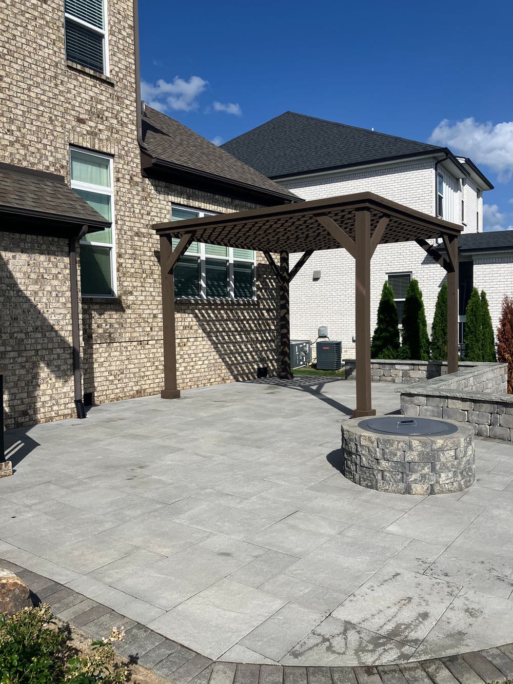 Patio area with wooden pergola and stone fire pit, surrounded by landscaped yard and homes.