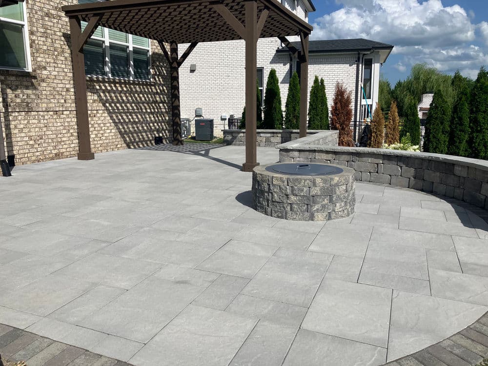Outdoor patio with stone flooring, fire pit, and wooden pergola under a blue sky.