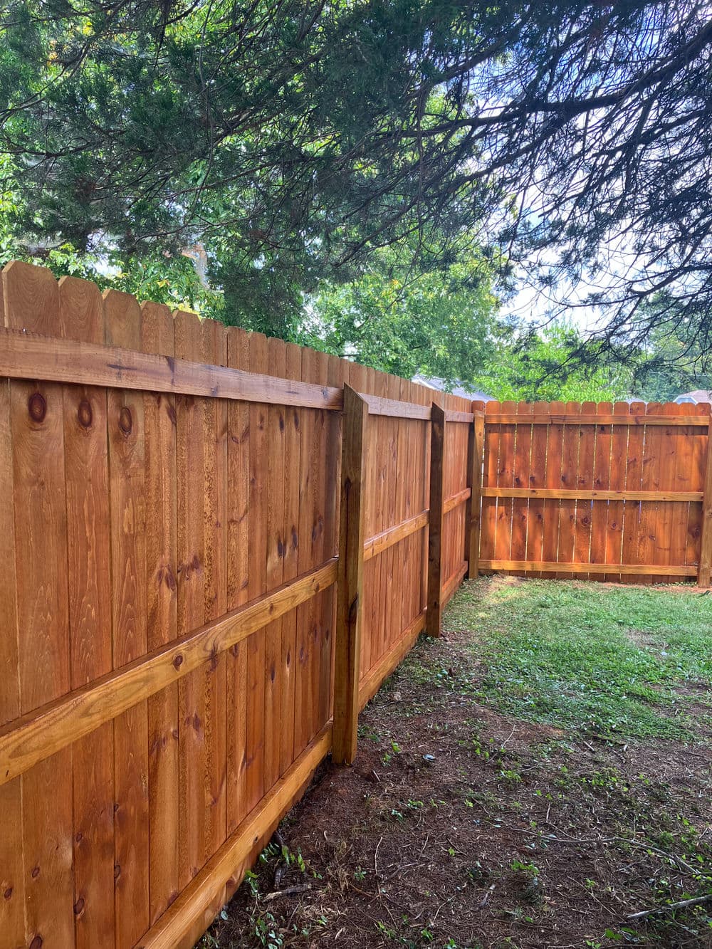 Newly installed wooden fence with a stained finish in a grassy yard surrounded by trees.