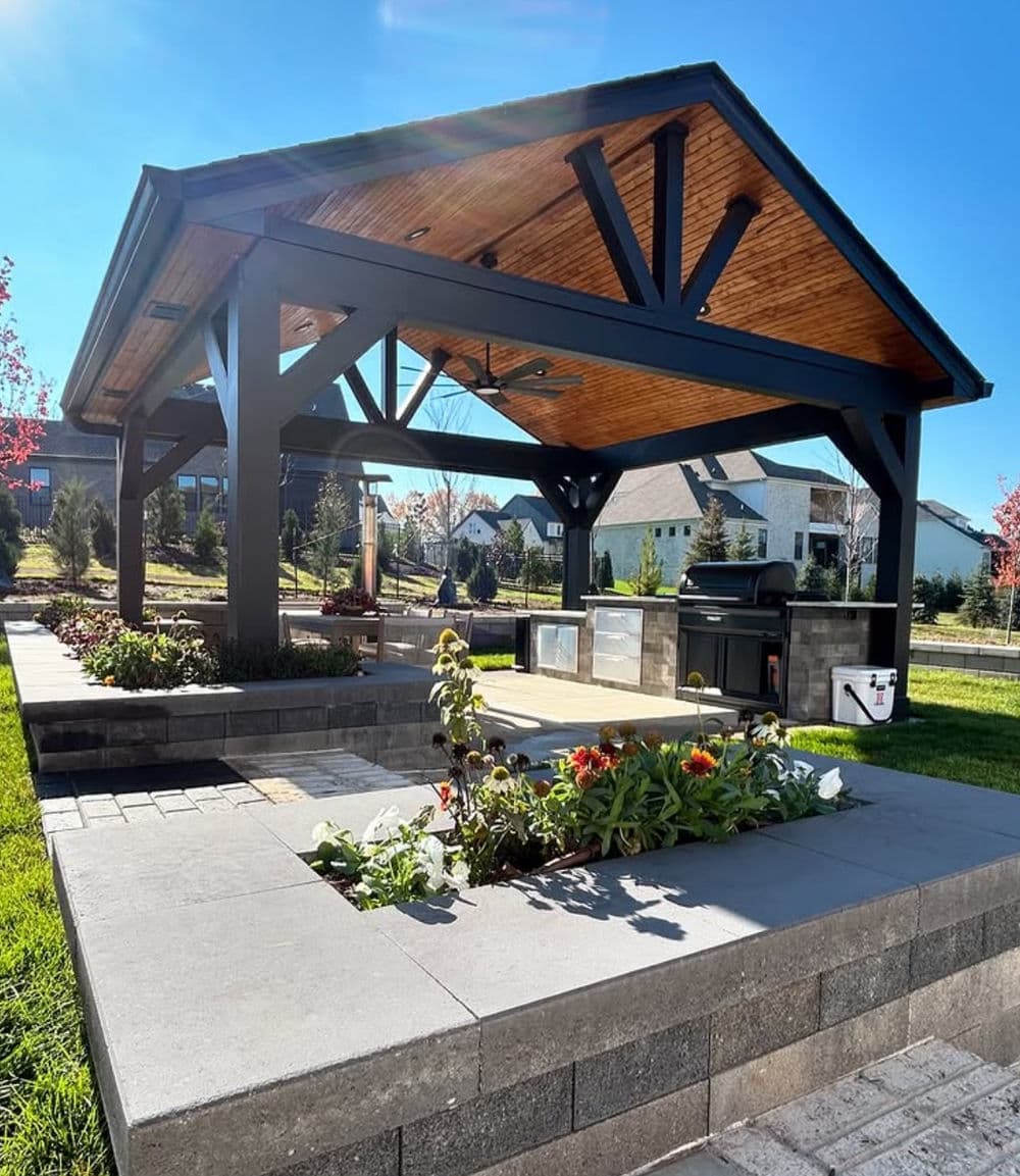 Outdoor kitchen gazebo with grill, seating, and landscaping in sunny backyard setting.
