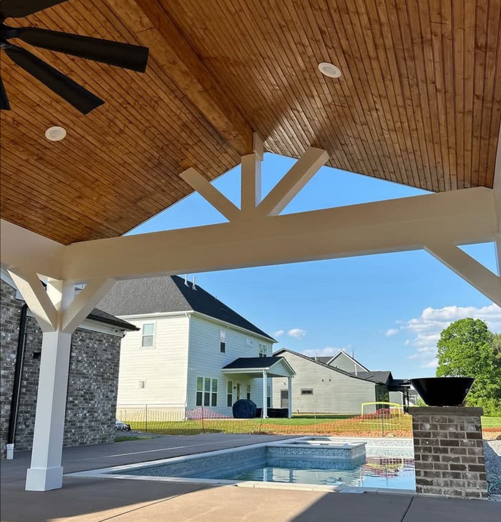 Covered patio with wooden ceiling, pool view, and houses in the background under clear blue sky.