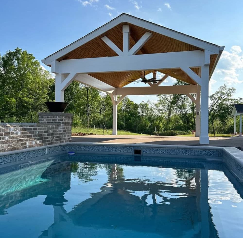Outdoor pool with a covered pavilion, surrounded by lush greenery under a clear blue sky.