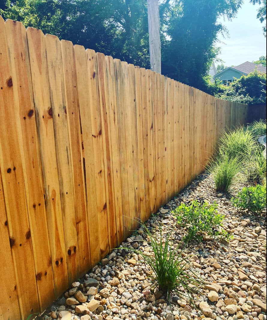 Wooden fence lining a garden with decorative stones and lush green plants. Bright sunlight.