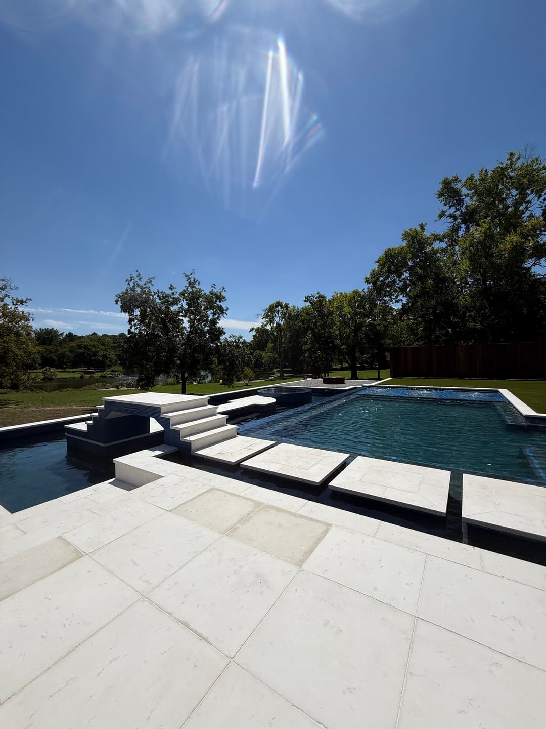 Modern outdoor pool with stone steps, surrounded by lush greenery under a clear blue sky.