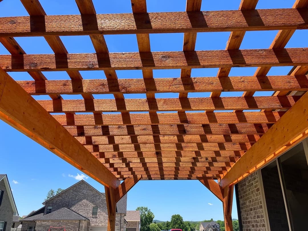 Wooden pergola structure viewed from below against a clear blue sky and houses in the background.