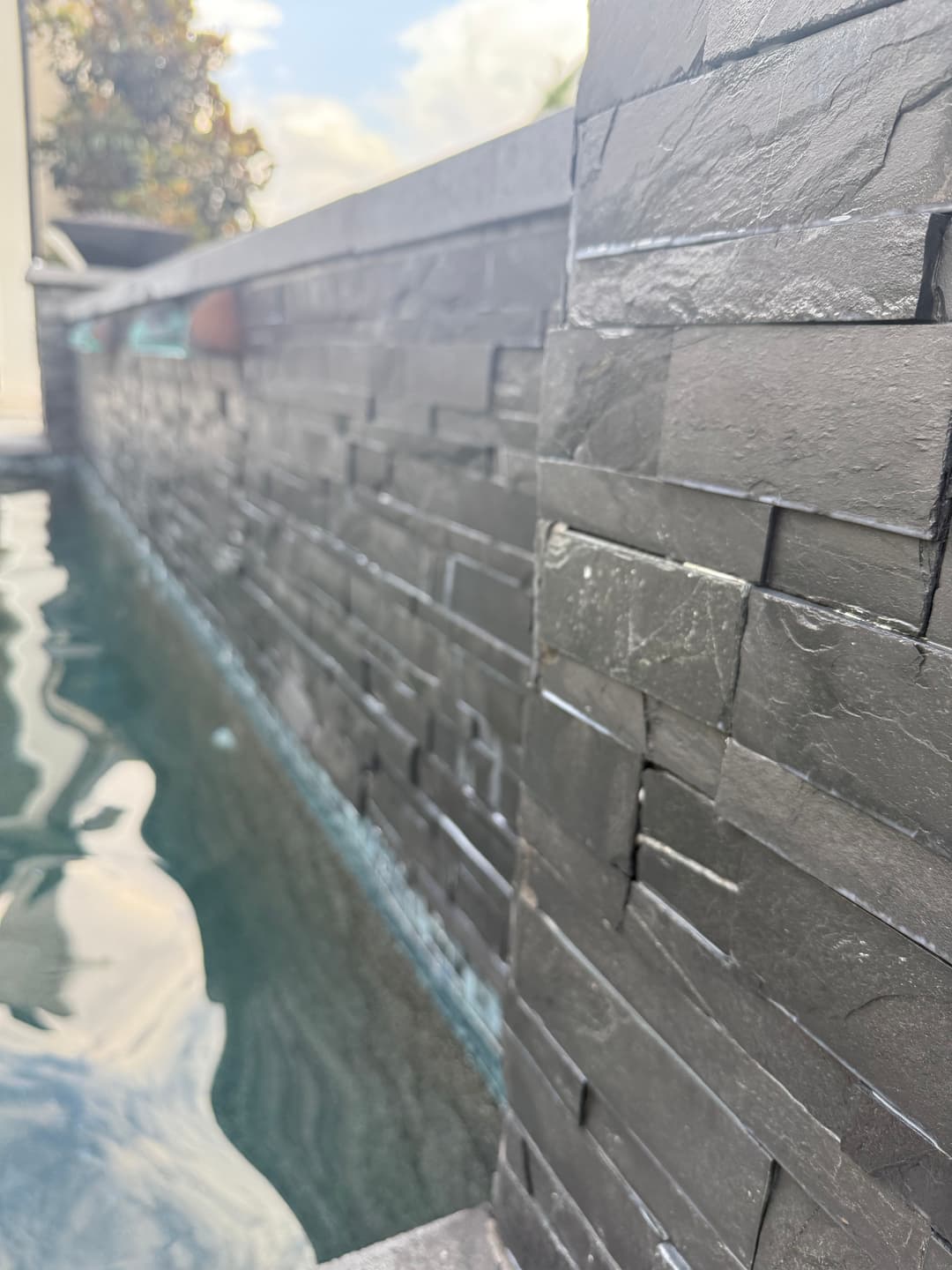 Close-up of textured stone wall beside a modern swimming pool, reflecting water and sky.