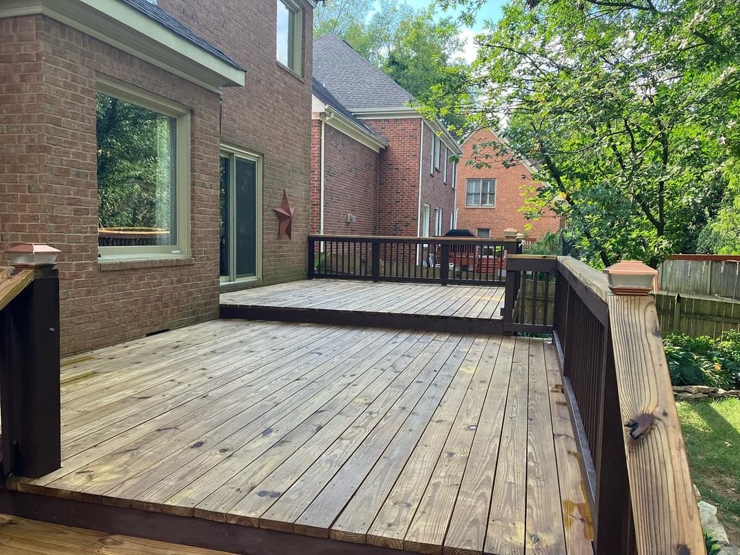 Spacious wooden deck attached to a brick house, surrounded by greenery and neighboring homes.