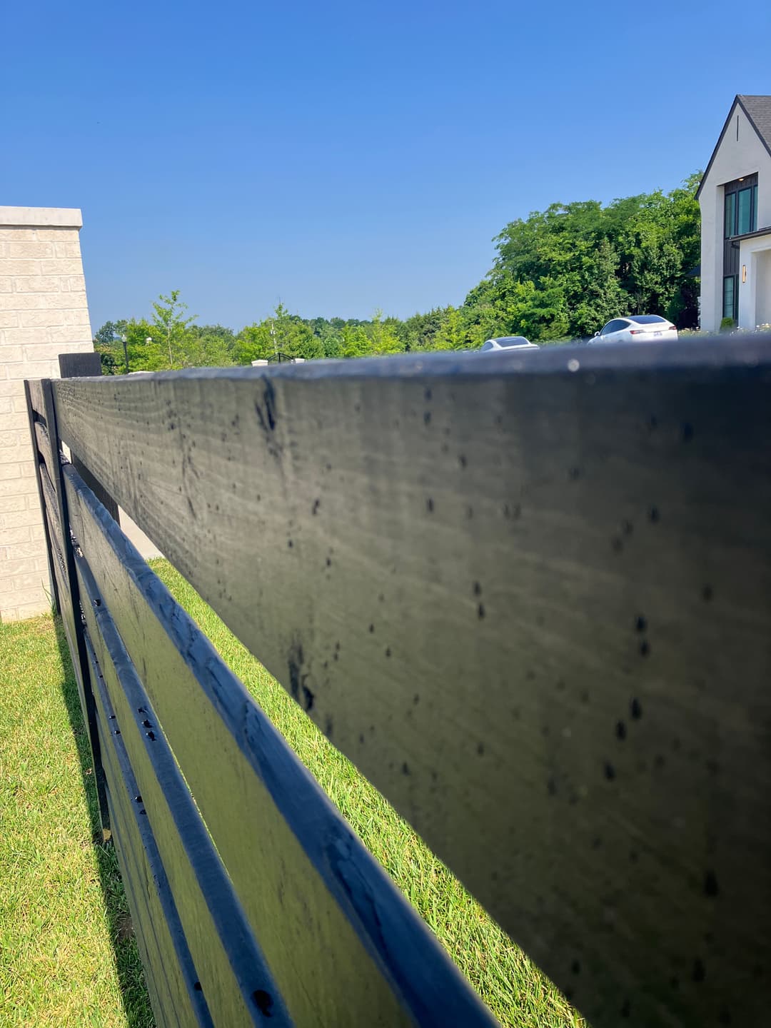 Black wooden fence with blue sky and greenery in the background, near a modern building.