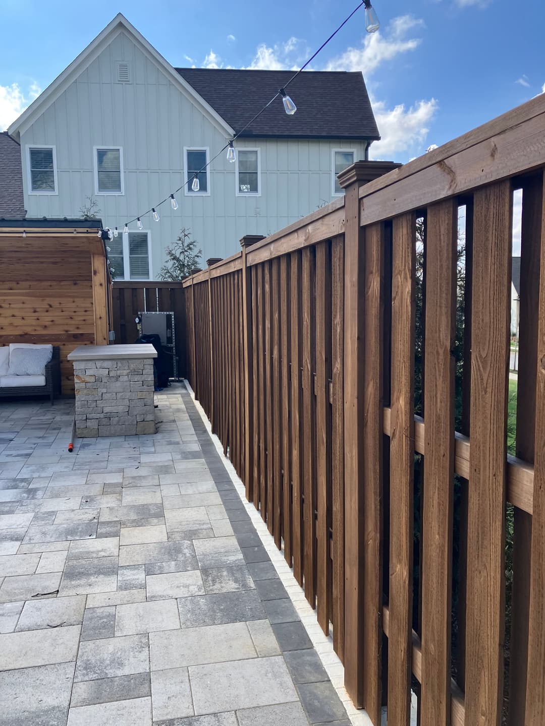 Wooden fence along a patio with pavers, outdoor seating, and string lights in residential setting.