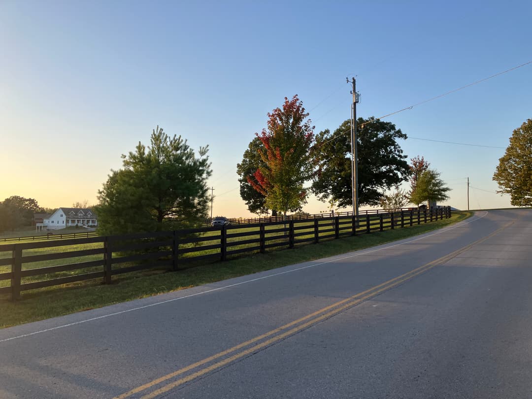 Scenic country road with black wooden fence, trees, and sunset sky in rural landscape.