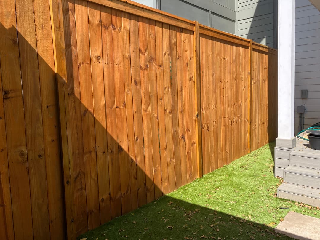 Wooden fence with natural finish, casting a shadow on green grass in a sunny backyard setting.