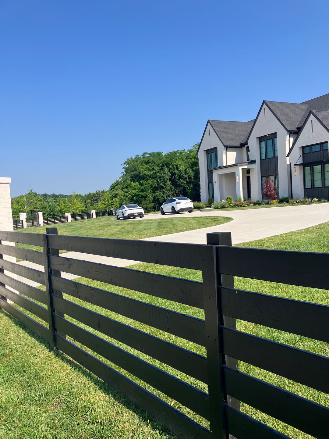 Modern house with two white cars parked, surrounded by a wooden fence and lush greenery.