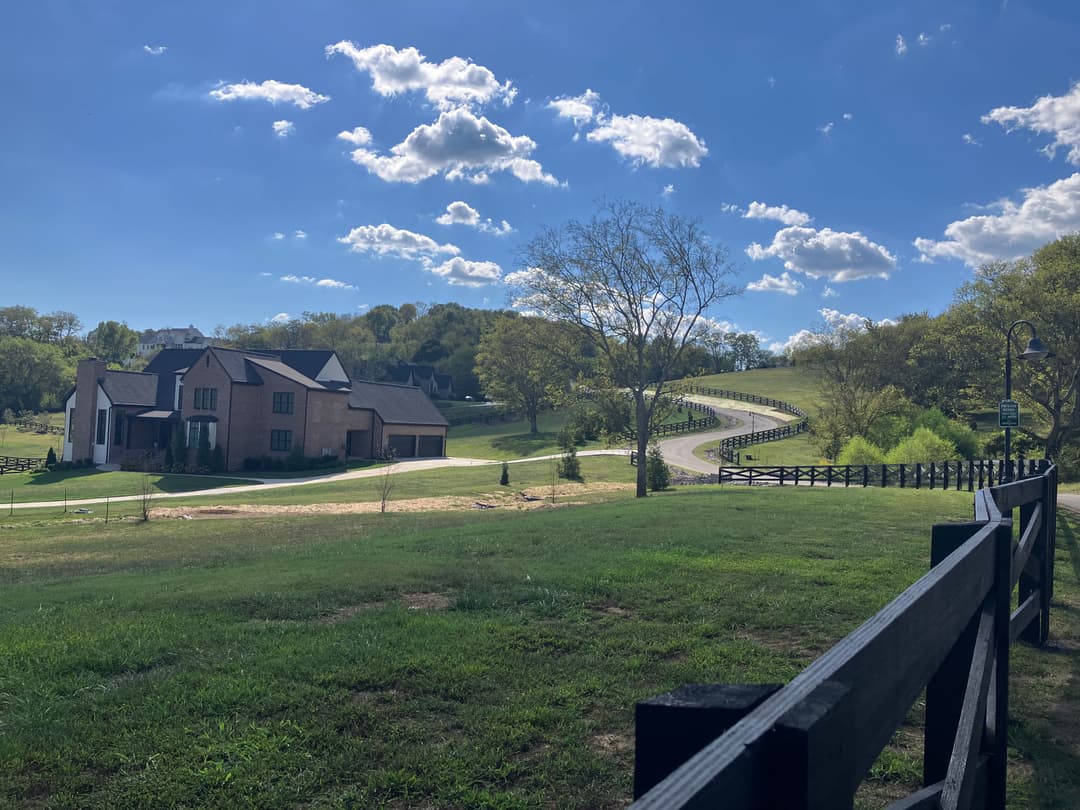 Scenic landscape featuring a country road, green fields, and a charming house under blue skies.