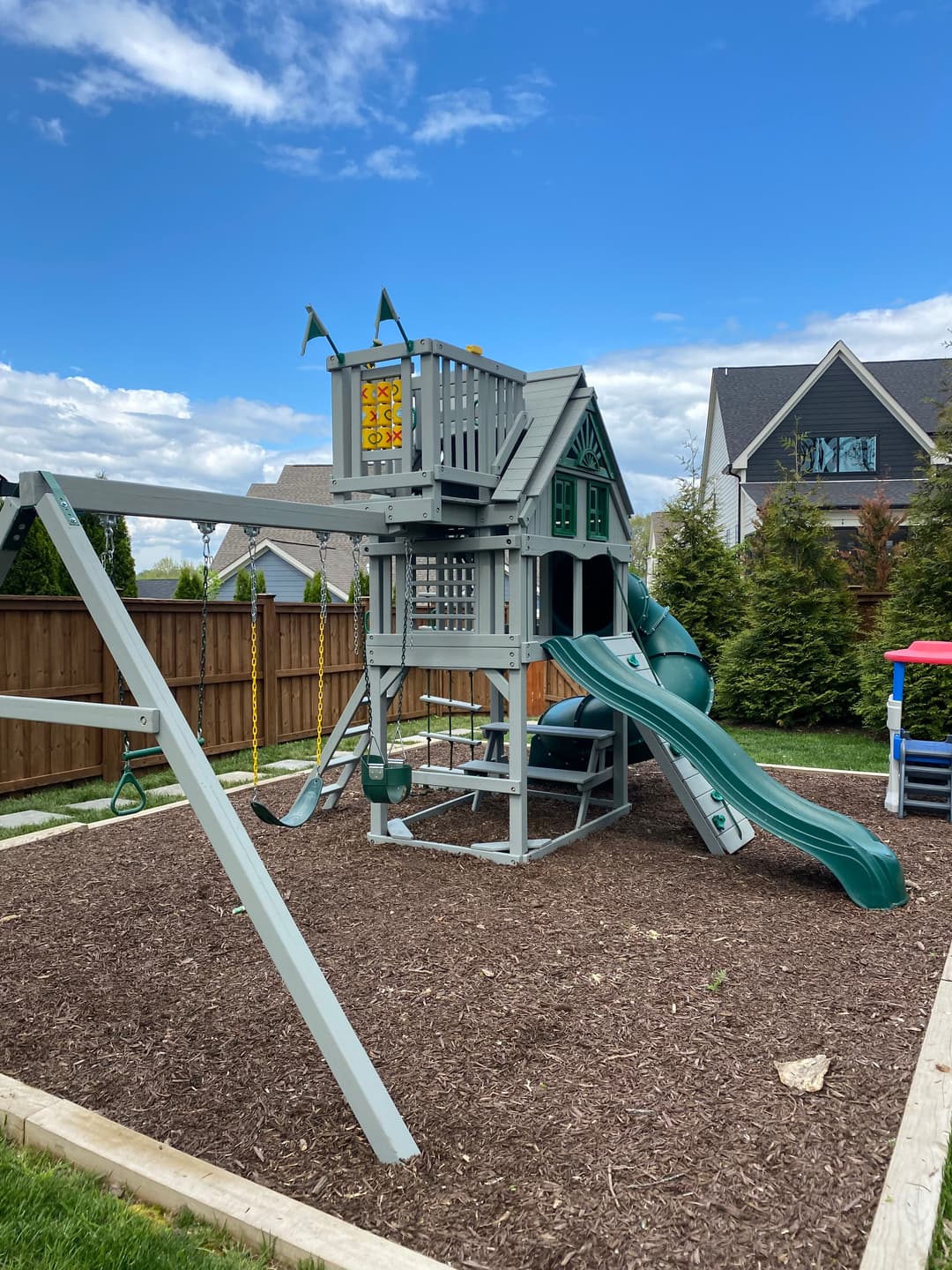 Backyard playground set with a slide, swings, and playhouse under a blue sky.