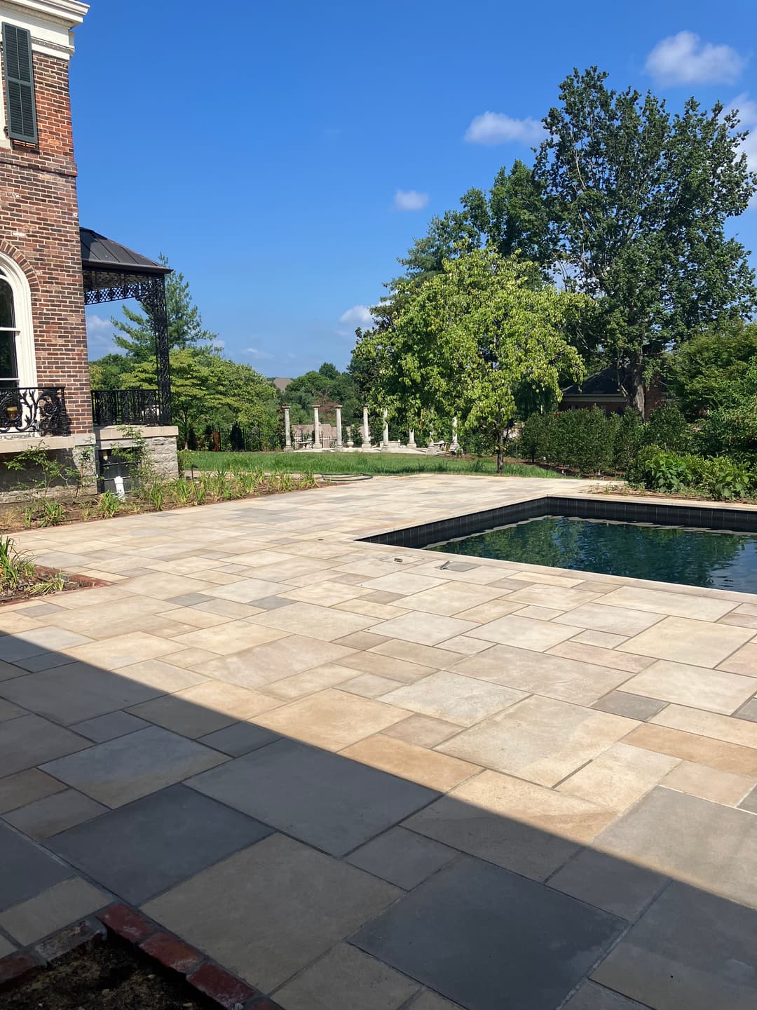 Elegant backyard with stone patio, swimming pool, and lush greenery under blue sky.