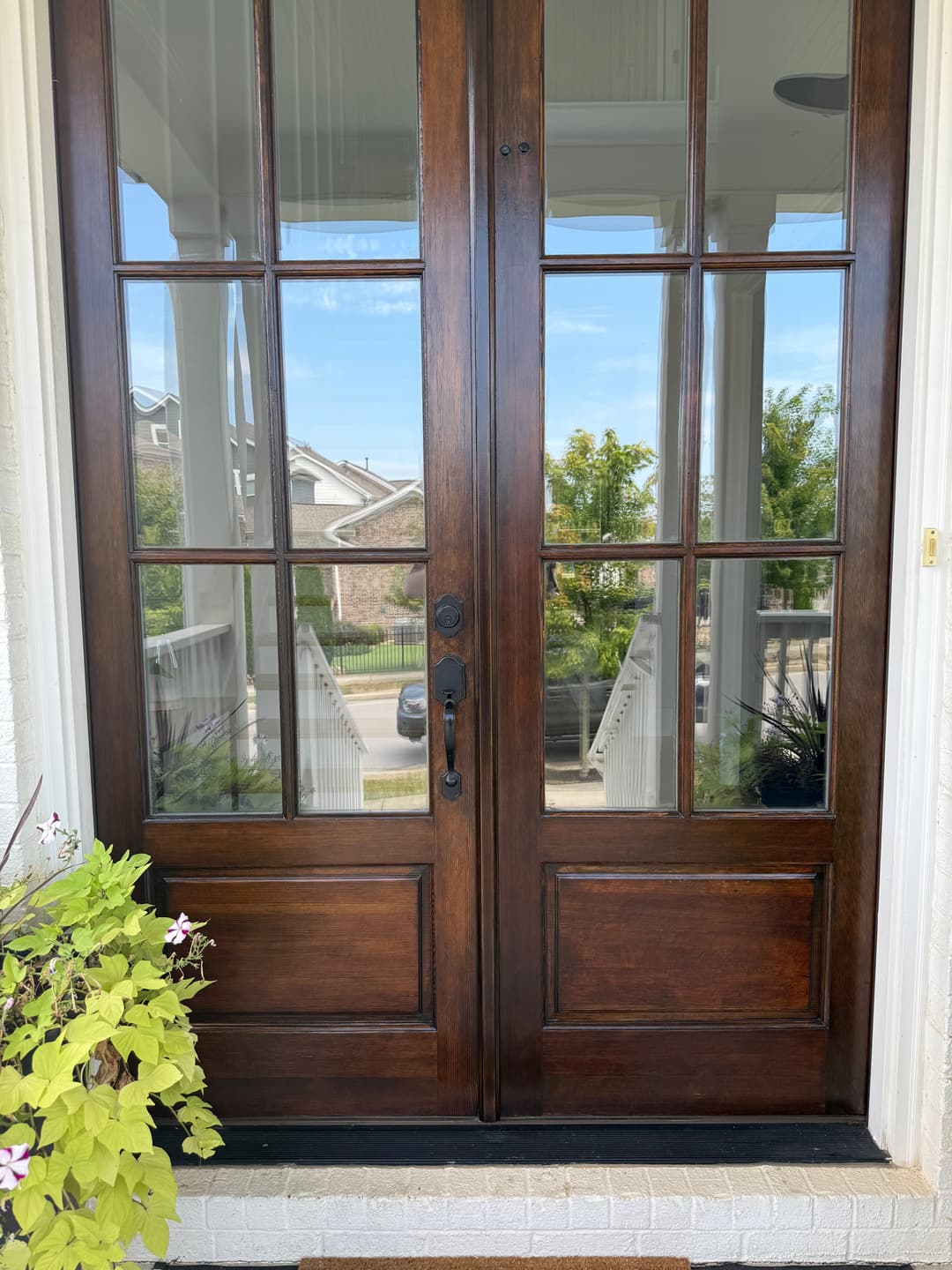 Elegant wooden double doors with glass panels, framed by a welcoming porch and greenery.