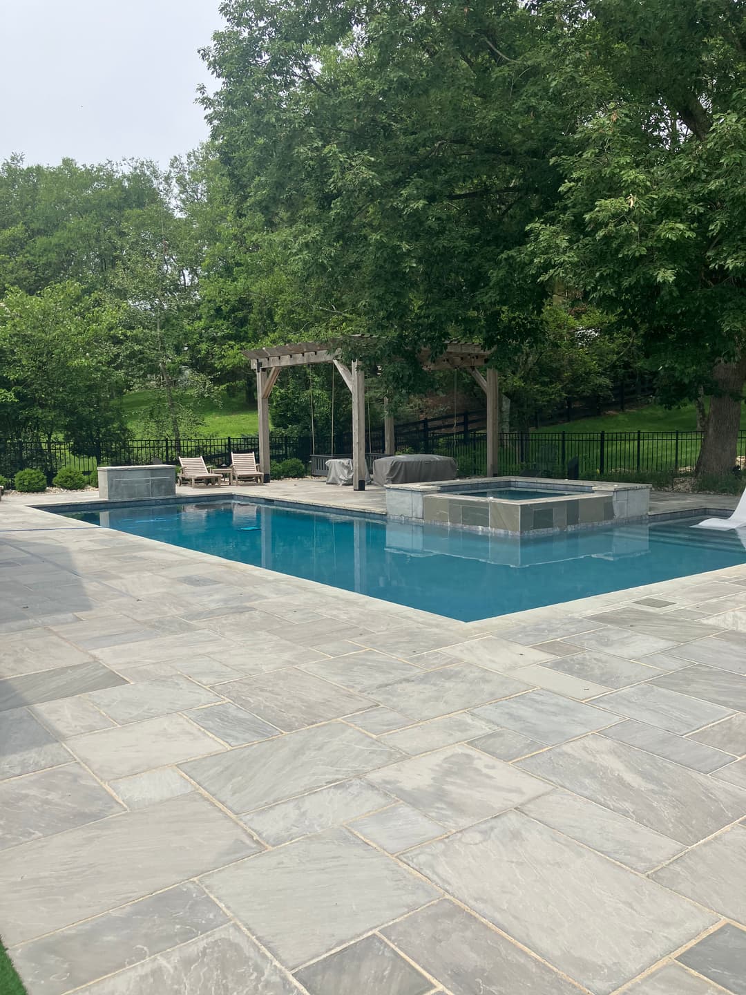 Modern backyard pool with stone patio, lounge chairs, and a pergola surrounded by greenery.
