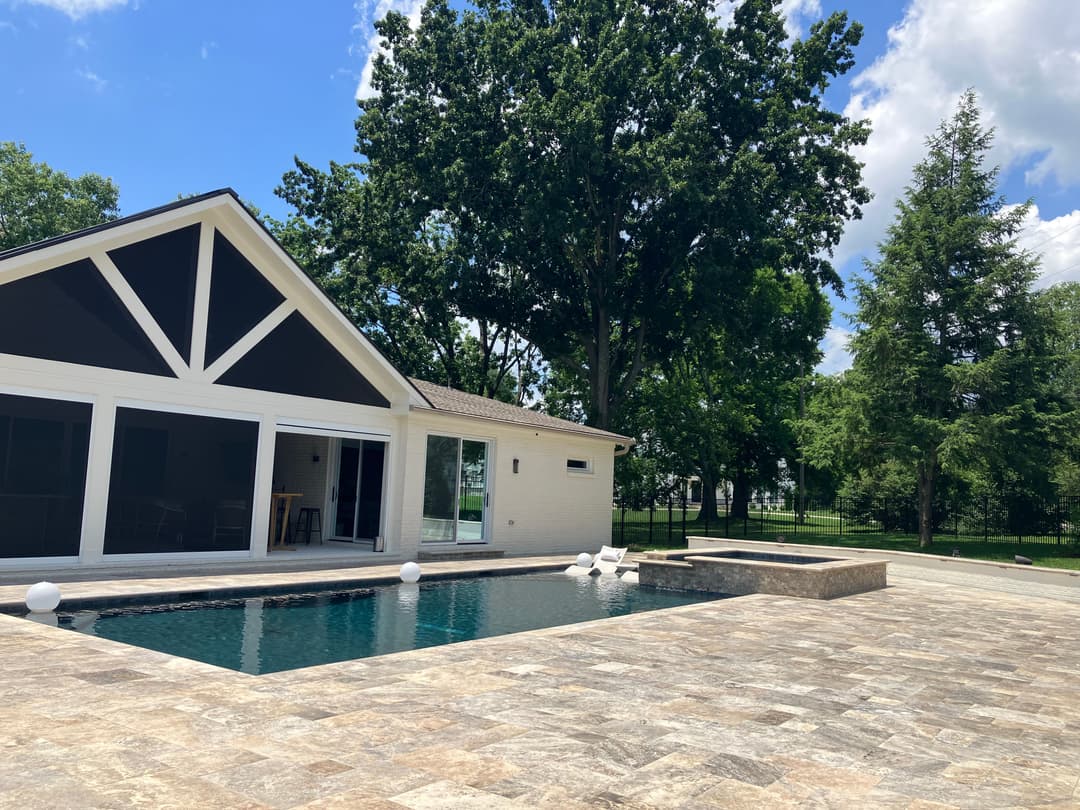 Modern house with pool and stone patio surrounded by lush green trees on a sunny day.