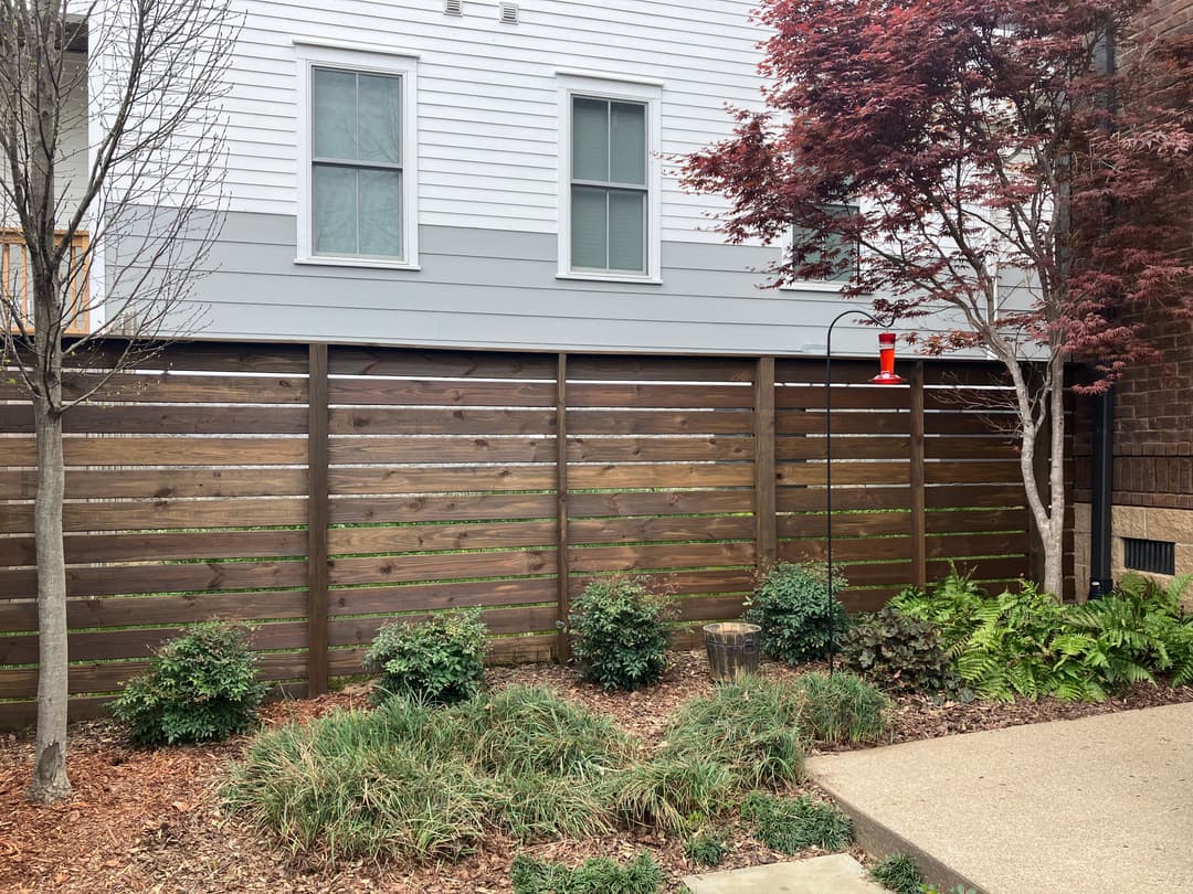 Wooden privacy fence with green shrubs, decorative plants, and a red bird feeder.