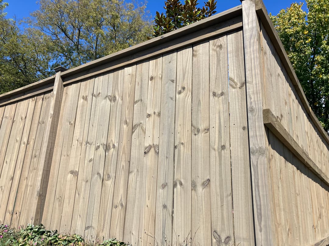Wooden fence with vertical planks against a blue sky and green trees.
