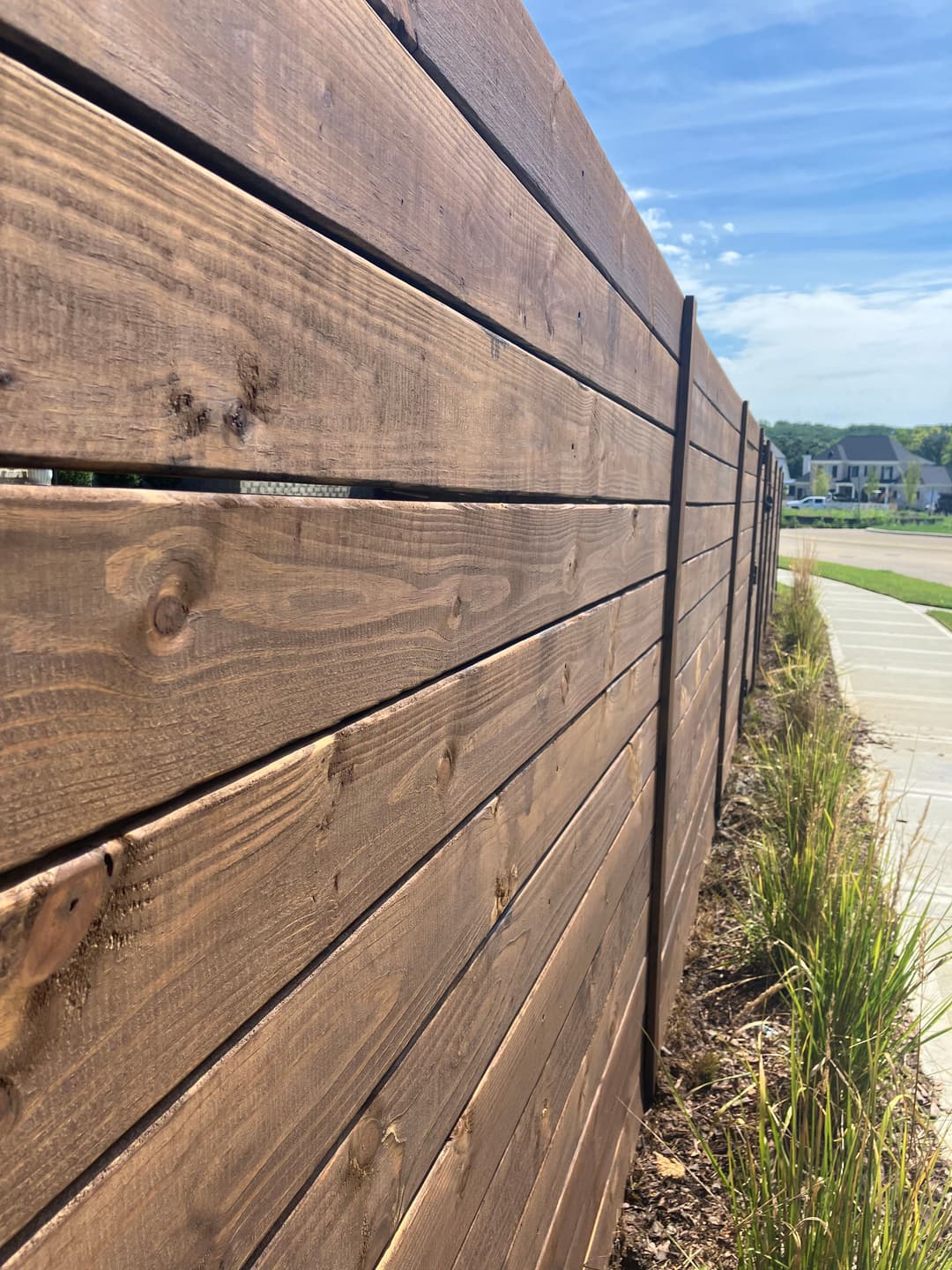 Wooden fence with a natural finish under a blue sky, bordered by greenery and a sidewalk.