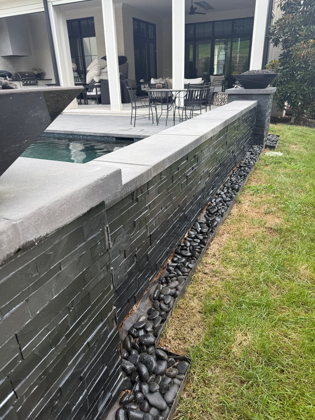 Modern backyard featuring a sleek stone wall, pool, and stylish dining area with black pebbles.