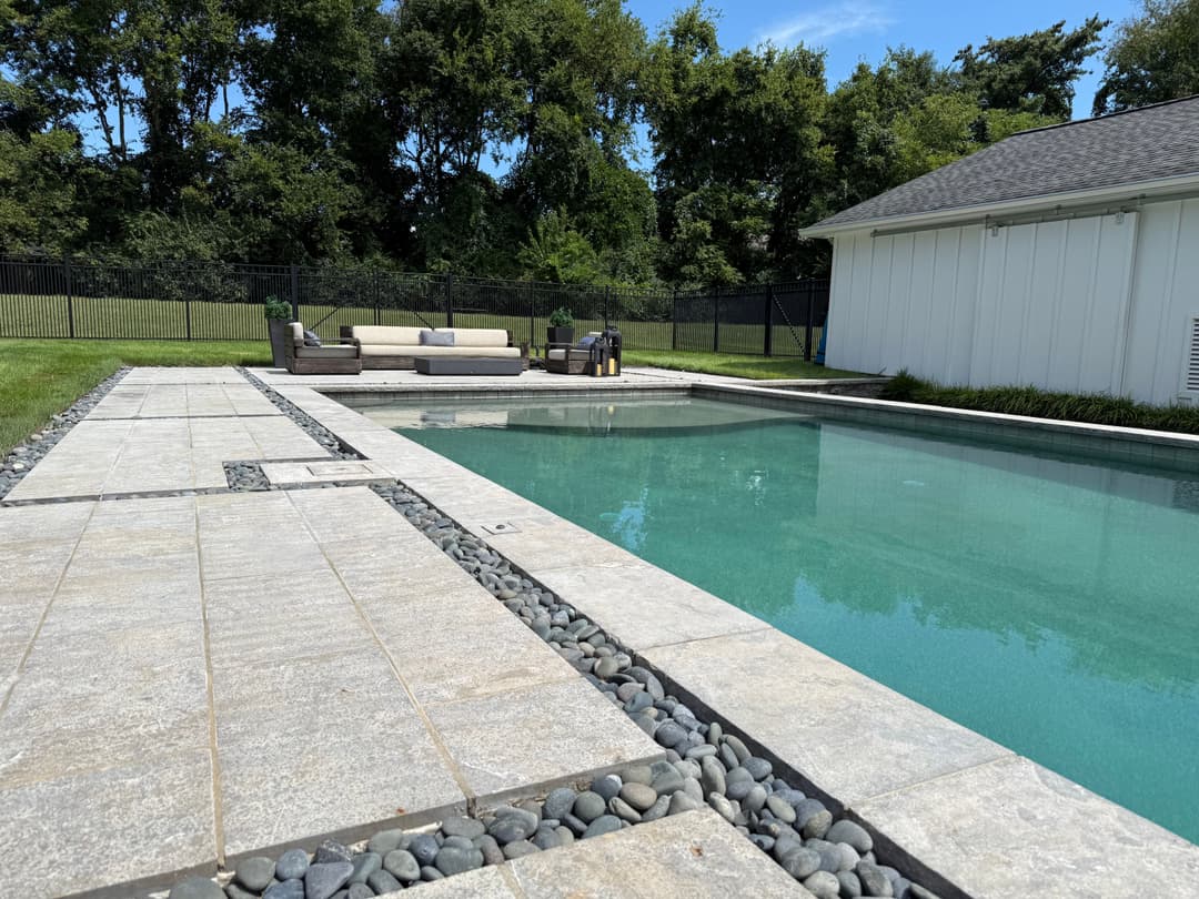 Modern backyard pool with stone patio and lounging area, surrounded by greenery.
